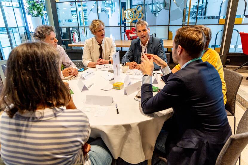 Zes mensen zitten aan een ronde tafel en praten met elkaar.